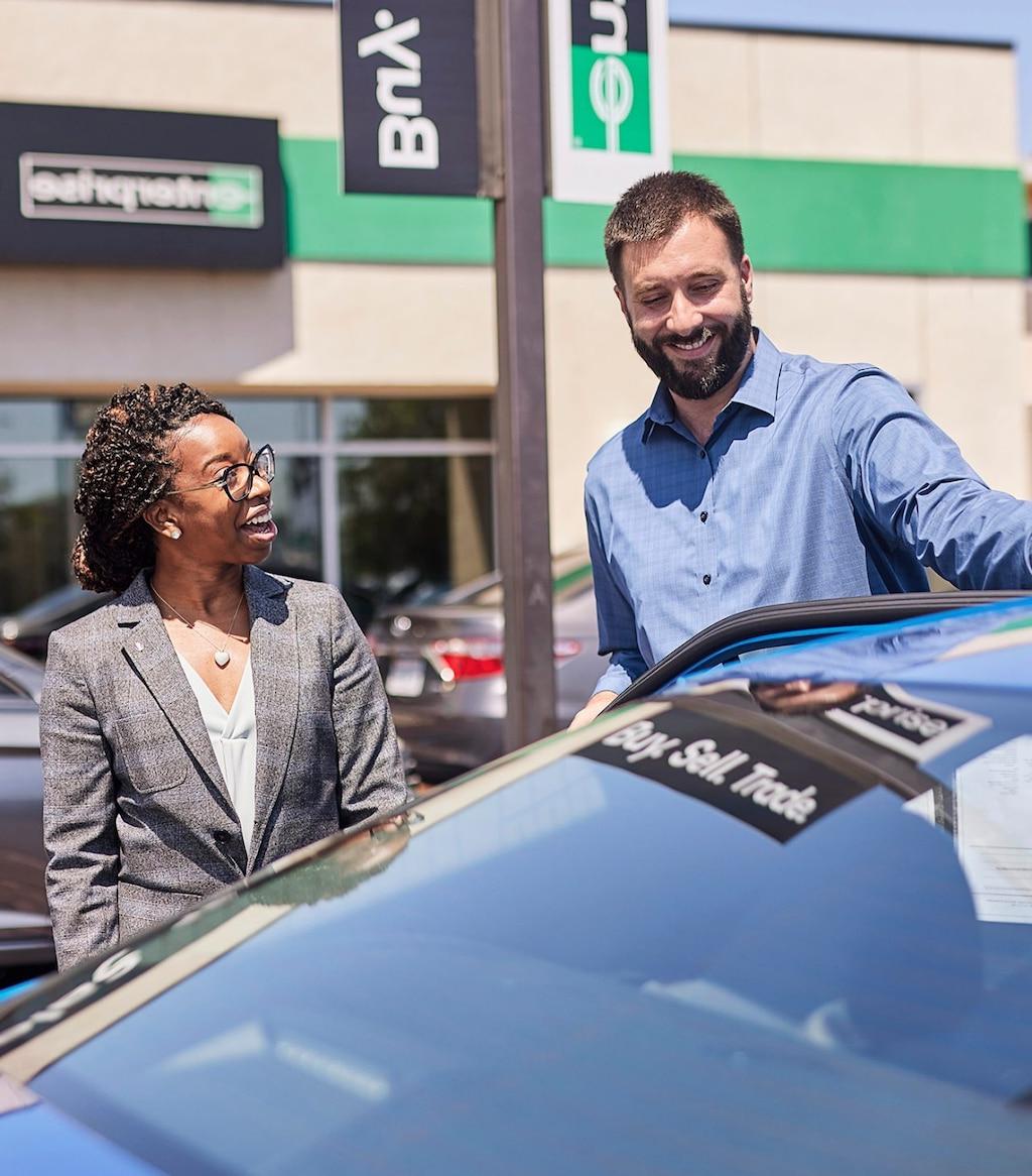 一位绅士站在一辆蓝色轿车前，与一位女士在企业汽车销售场上交谈.